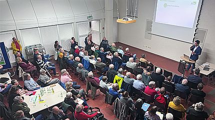 Eine Präsentation in einem Seminarraum mit vielen Teilnehmern, die auf Stühlen sitzen und einem Redner zuhören, der an einem Pult steht und eine Präsentationsfolie zeigt.