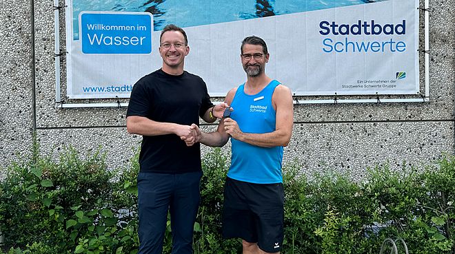 Zwei Personen vor dem Stadtbad Schwerte. Sie geben sich die Hände und Florian Grojer übergibt eine KI-Uhr Anden ehemaligen Schwimmbadleiter Thomas Jeserig.