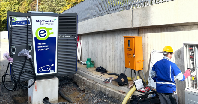 Eine neue Elektroauto-Ladestation von den Stadtwerken Schwerte mit 300 kW Leistung wird installiert, daneben ein Arbeiter mit Schutzhelm und Arbeitskleidung an einem geöffneten Stromkasten