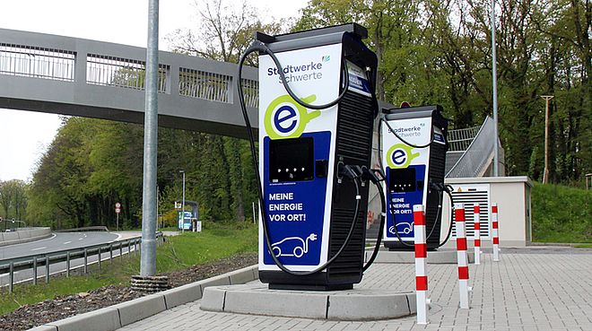Zwei Ladesäulen der Stadtwerke Schwerte mit der Information "Mehr Energie vor Ort". Die Ladesäulen und dazugehörigen Parkplätze befinden sich neben einer Schnellstraße und einer Fußgängerbrücke