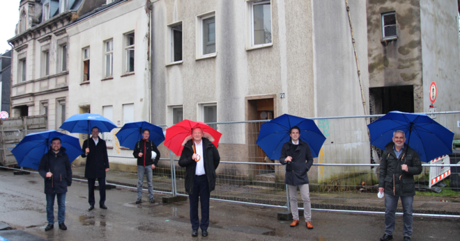 Sechs Männer mit geöffneten Schirmen bei Regen vor einem unbewohnten, etwas heruntergekommenen Haus