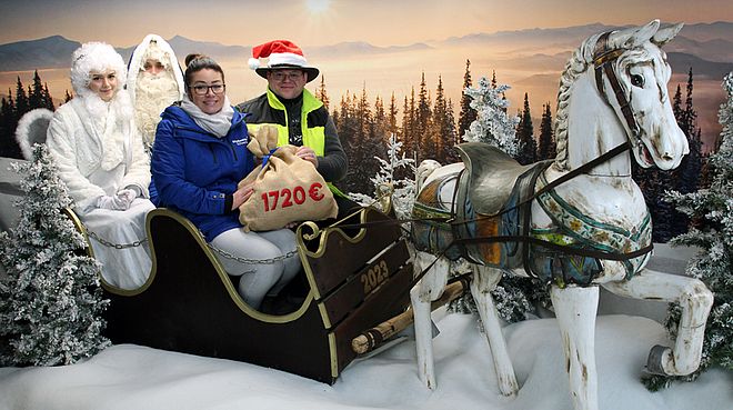 Vier Personen sitzen in einem Schlitten, der von einem weißen Pferd gezogen wird, vor einer Winterlandschaft mit verschneiten Bäumen und Bergen im Hintergrund. Eine Person im Schlitten hält einen Sack mit der Aufschrift "1720 €".