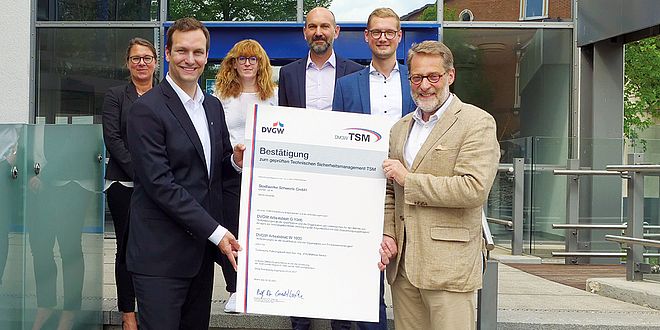 Sebastian Kirchmann, Geschäftsführer der Stadtwerke Schwerte, und Dr. Bernhard Klocke vom DVGW halten gemeinsam das Zertifikat "Bestätigung zum geprüften Technischen Sicherheitsmanagement TSM" hoch, im Hintergrund vier weitere Mitarbeiter der Stadtwerke Schwerte