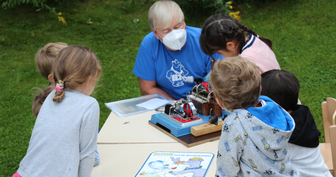 5 kleine Kinder und ein Mann mit Maske,  betrachten auf einer Wiese an einem Tisch ein Modell zum Thema Energie