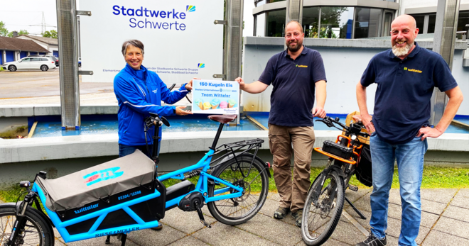 Eine Frau und zwei Männer stehen mit einem Fahrrad vor dem Verwaltungsgebäude der Stadtwerke Schwerte, die Frau überreicht symbolisch einen Gutschein für 150 Kugeln Eis