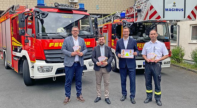 Vier Männer vor zwei Fahrzeugen der Feuerwehr mit Broschüren in der Hand