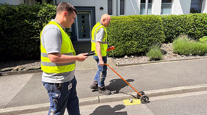 Zwei Männer mit gelben Warnwesten schieben eine Teppichsonde über den Asphalt, um Gasleitungen auf mögliche Fehler zu überprüfen.