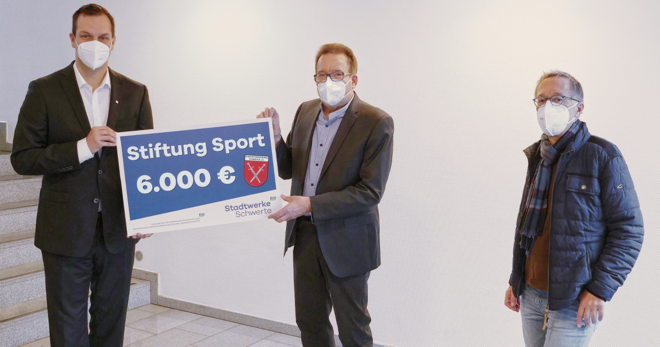 Drei Personen mit Maske vor einer Treppe im Hausflur, Sebastian Kirchmann, Geschäftsführer der Stadtwerke Schwerte, überreicht überreicht einen symbolischen Check mit dem Schriftzug "Stiftung Sport 6.000 €"
