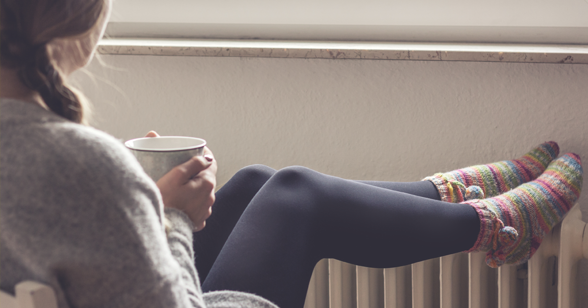 Eine Frau sitzt vor einem Fenster, die Füße auf der Heizung. In der Hand hält sie mit beiden Händen eine Tasse. 
