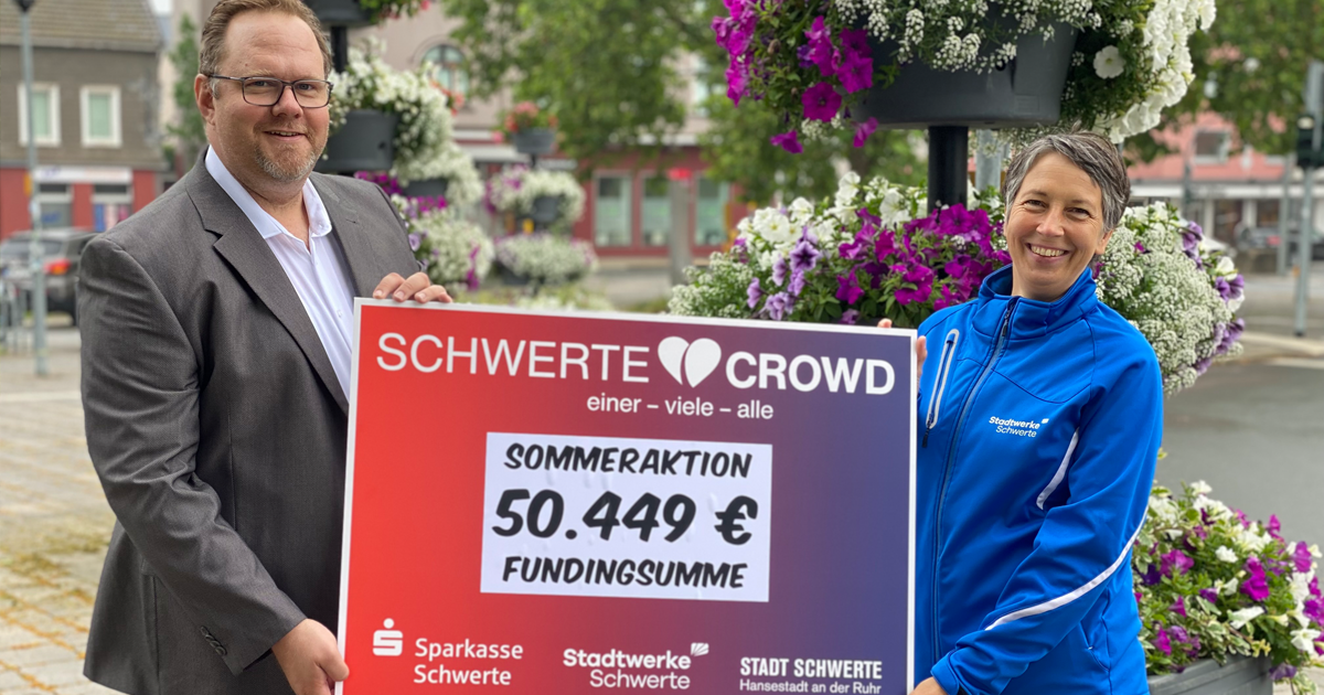 Die Mitarbeiterin der Stadtwerke Schwerte übergibt einer weitere Personen einen symbolischen Check mit dem Schriftzug "Schwerte Crowd, Sommeraktion 50.499 € Fundingsumme"