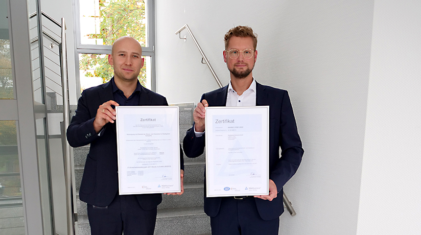Zwei Mitarbeiter der Stadtwerke Schwerte stehen im Flur eines Gebäudes und halten jeweils ein eingerahmtes Zertifikat in den Händen.