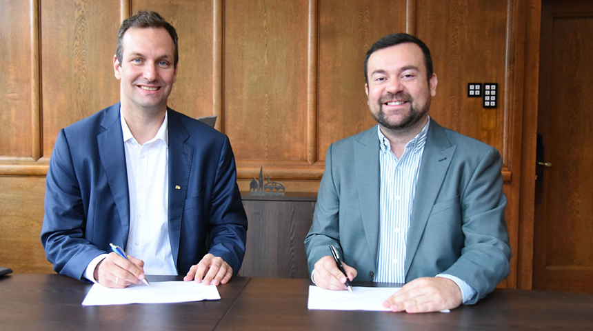 Geschäftsführer der Stadtwerke Schwerte, Sebastian Kirchmann, und Bürgermeister Dimitrios Axourgos sitzen an einem Holztisch und unterzeichnen Dokumente, sie lächeln in die Kamera.