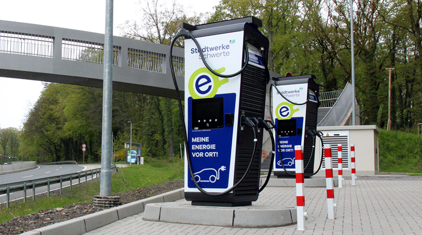 Zwei Ladesäulen der Stadtwerke Schwerte mit der Information "Mehr Energie vor Ort". Die Ladesäulen und dazugehörigen Parkplätze befinden sich neben einer Schnellstraße und einer Fußgängerbrücke