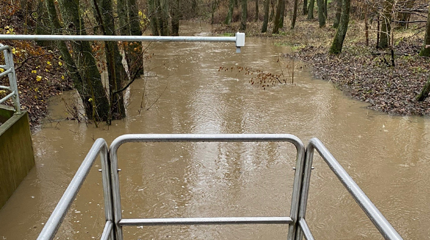 Ein überfluteter Bachlauf in einem Waldgebiet, davor eine Art Aussichtsplattform und darüber das LoRaWAN, ein Hochwasserfrühwarnsystem
