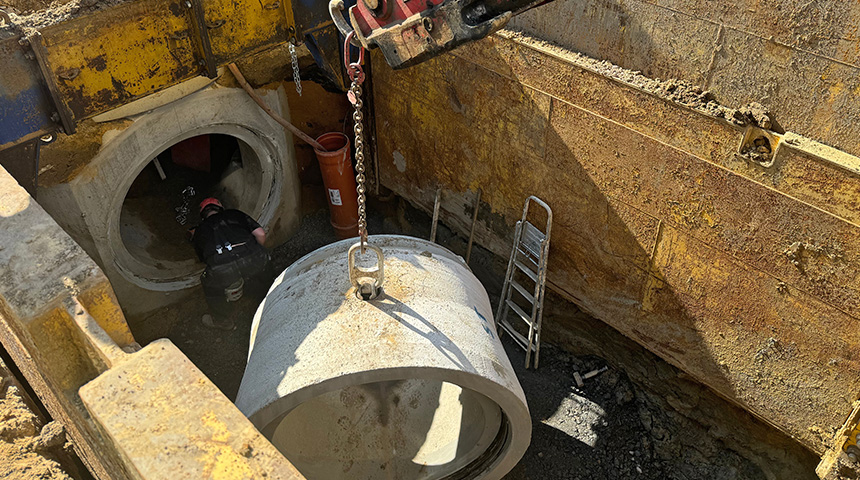 Arbeiter in Schutzkleidung arbeitet in einer Baugrube neben einem großen Betonrohr, das mit einem Kran herabgelassen wird