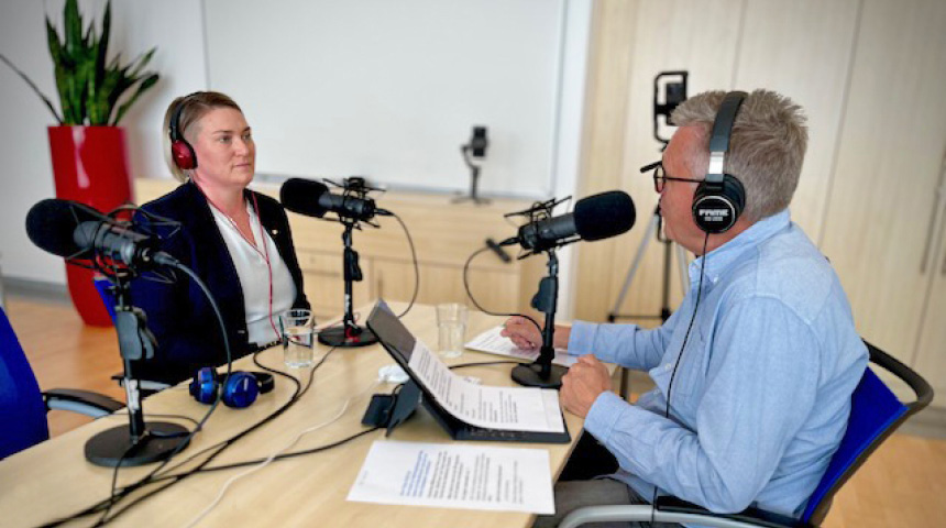 Zwei Personen führen ein Podcast-Interview mit Mikrofonen und Kopfhörern an einem Tisch, Notizen liegen vor ihnen.