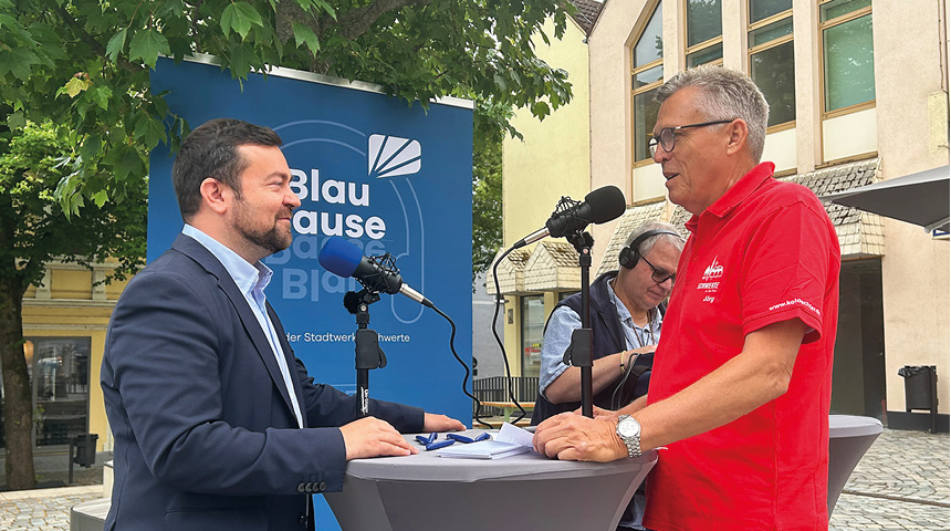 Moderator Jörg Przystow und Bürgermeister Dimitrios Axourgos führen ein Podcast-Interview an einem Stehtisch mit zwei Mikrofonen und Notizen. Im Hintergrund ein Baum und ein blaues Plakat mit dem Schriftzug "BlauPause".
