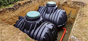 Zwei schwarze Kunststoff-Wasserzisternen mit grünen Deckeln in einer Erdgrube, umgeben von Erde und einem roten Rohr