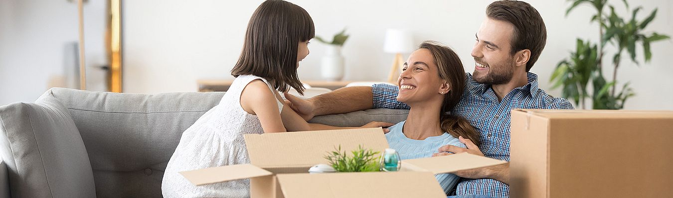 Familie mit zwei Erwachsenen und einem Kind sitzt auf einem Sofa, umgeben von Umzugskartons als Symbolbild für die Umzugsplanung.
