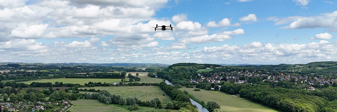 Eine Drohne fliegt über eine grüne Landschaft in Schwerte, mit Wiesen, Bäumen, Flusslauf und Wohnhäusern im Hintergrund unter einem blauen Himmel mit weißen Wolken.