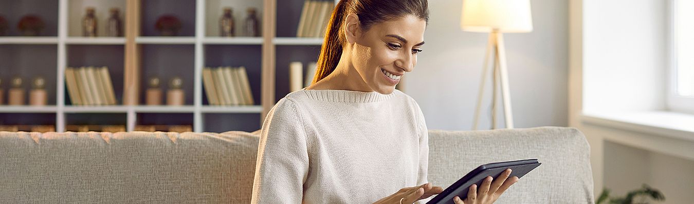 Eine Frau sitz lächelnd auf einem Sofa und hält ein Tablet in ihren Händen. Im Hintergrund sind Bücherregale und eine Stehlampe.