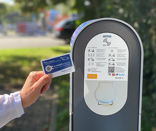 Eine Hand hält die Ladekarte der Stadtwerke Schwerte an eine Ladesäule. Oben rechts auf der Karte ist ein Elektroauto als Umriss zu sehen